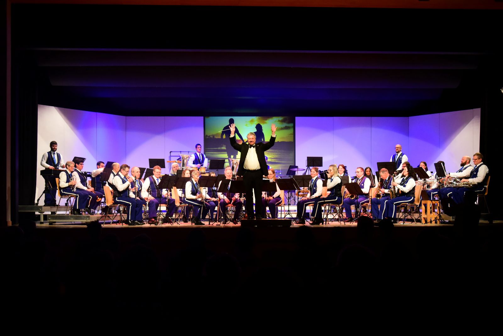 Die Harmoniemusik Schwanden steht in Uniform auf der Bühne und schaut ins Publikum, während der Dirigent im schwarzen Anzug den Zuschauern winkt